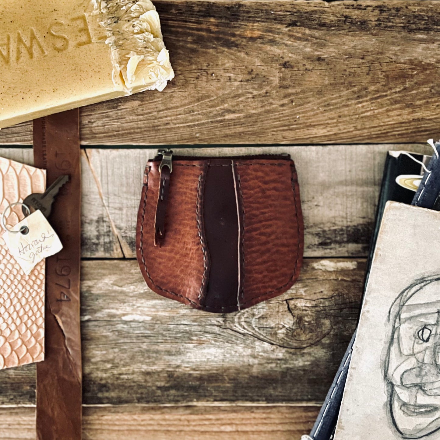 image of Sondera 1974s Little River Coin Pouch in Hickory and Chestnut leather resting on a rustic wooden backdrop with beeswax, leather strips and illustrations are surrounding the pouch.