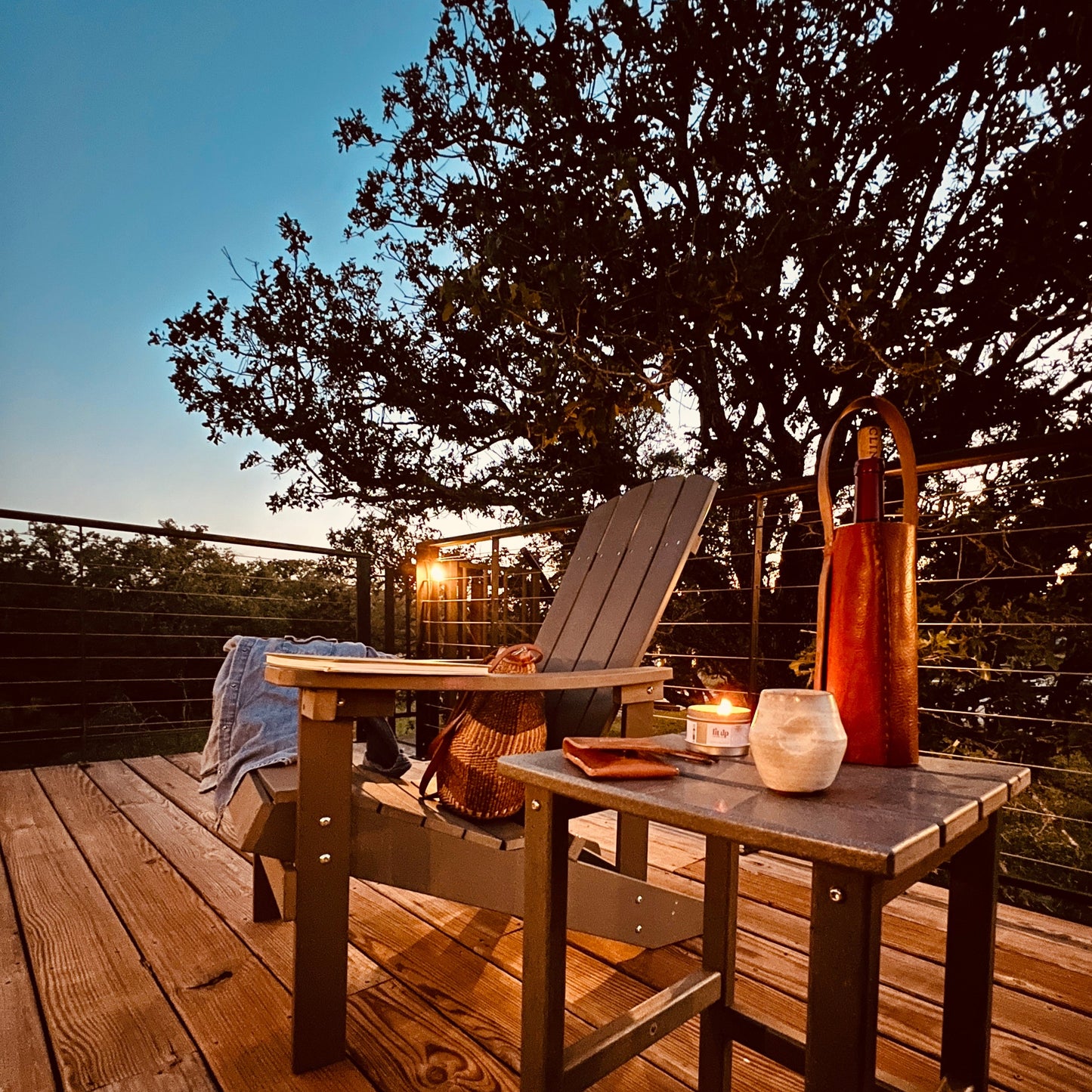 Wooden deck with outdoor furniture, candles, and a leather wine carrier under a tree at dusk.