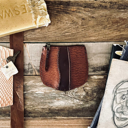 image of Sondera 1974s Little River Coin Pouch in Hickory and Chestnut leather resting on a rustic wooden backdrop with beeswax, leather strips and illustrations are surrounding the pouch.