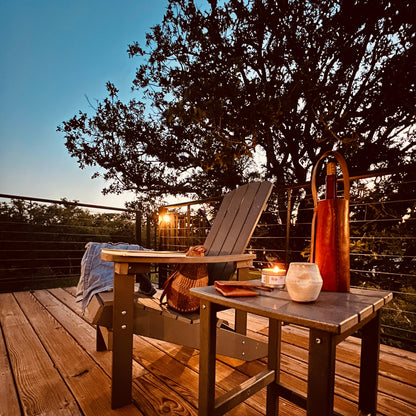 Wooden deck with outdoor furniture, candles, and a leather wine carrier under a tree at dusk.