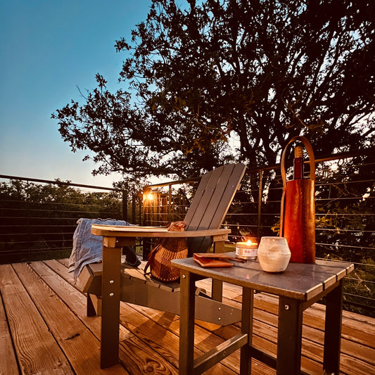Wooden deck with outdoor furniture, candles, and a leather wine carrier under a tree at dusk.